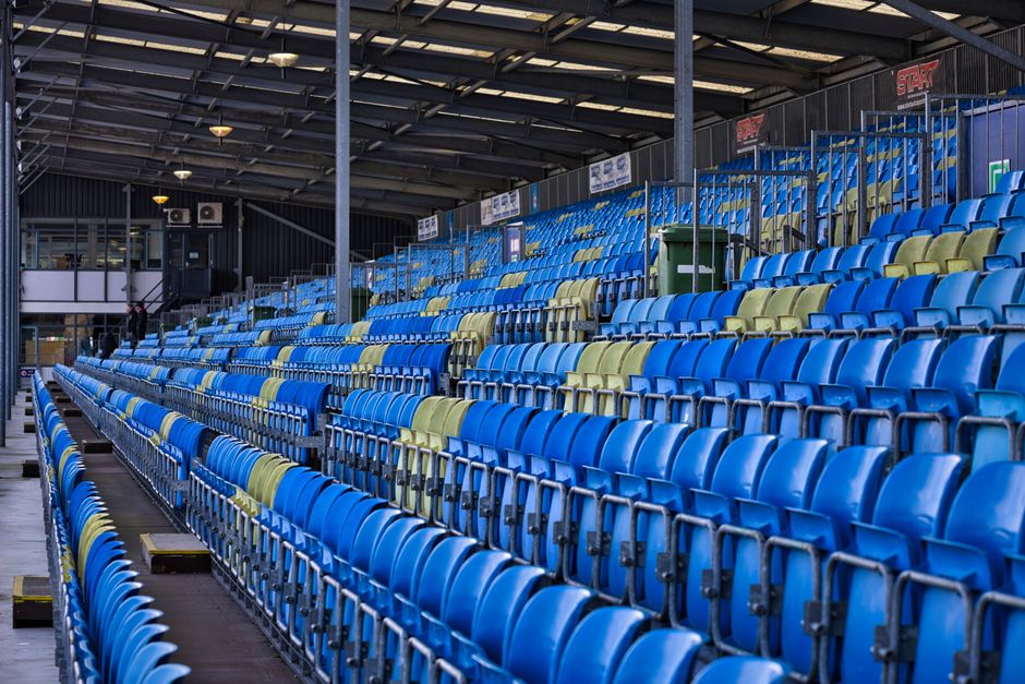 Empty red plastic stadium seats arranged in rows within a professional sports arena, with a concrete walkway running between the seats and towards the upper tiers. The seating area is illuminated by n